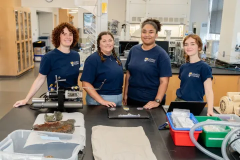 Jill McDermott, Jessica Rodgers, Jada Siverand and Esmira Bibaj pose for a photo in a lab with water testing equipment in front of them.